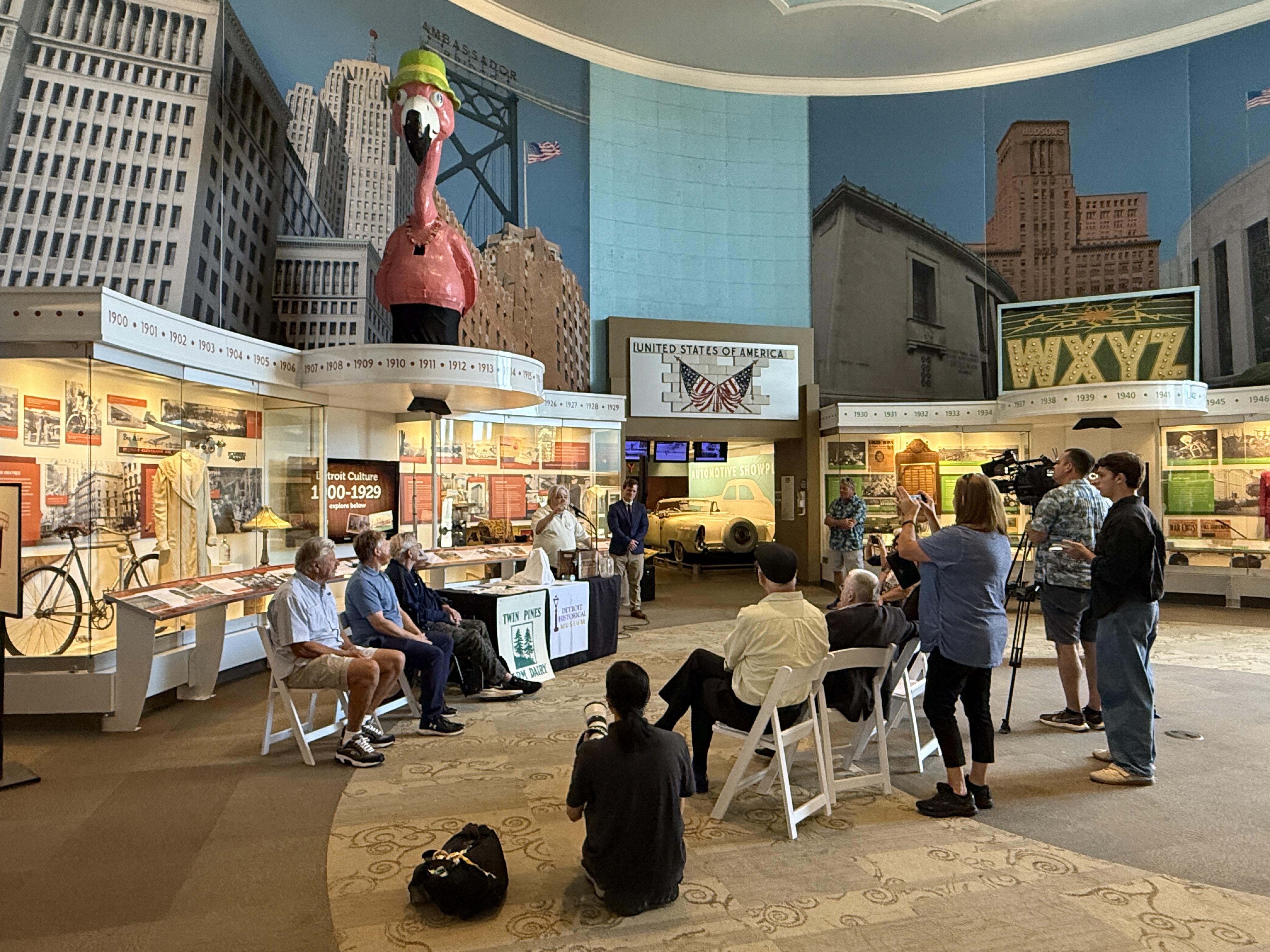 People seated and standing in a museum exhibit room with a large flamingo statue and city skyline backdrop.