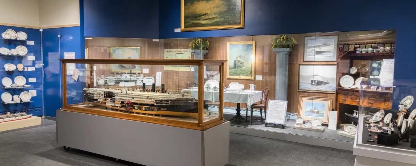Museum exhibit with a ship model in a glass case, surrounded by maritime paintings and artifacts on the wall.