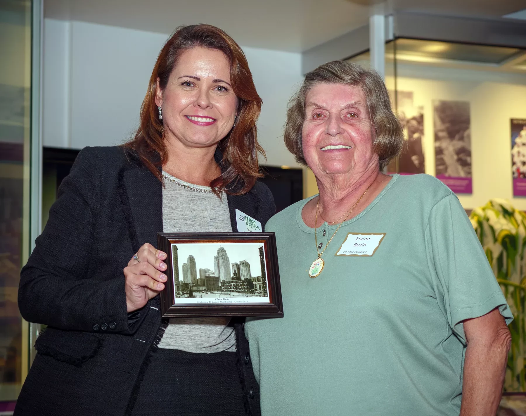 Two women smiling, one holding a framed photo of a cityscape, standing indoors.