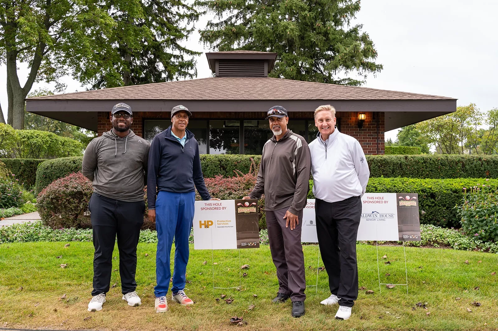 Four men in athletic gear standing in front of a building and hedge, next to a sign that says this hole sponsored by H&P Protective Services, Detroit Historical Scociety History of Golf Classic.