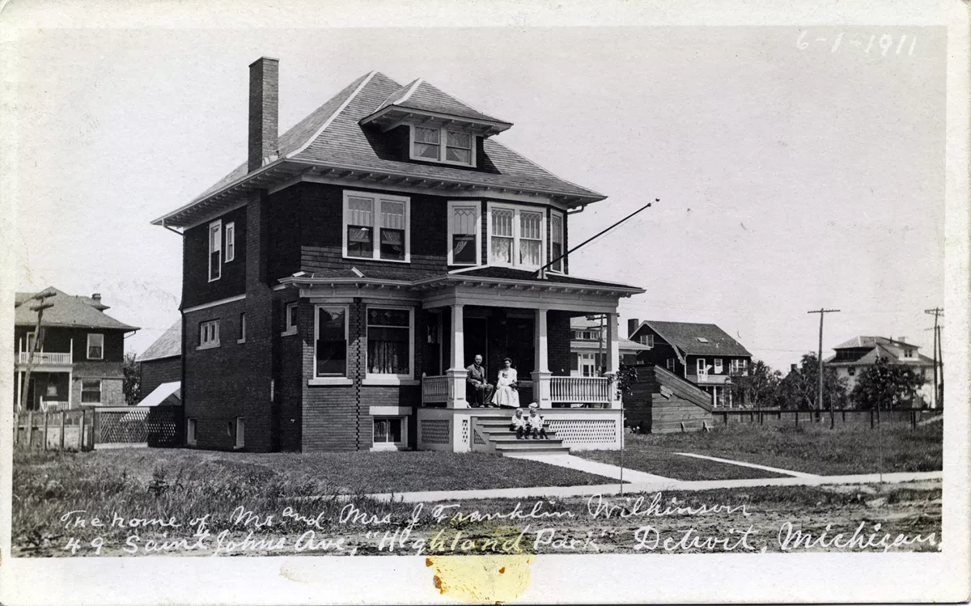 Black and white photo of a two-story house with people on the porch, surrounded by a grassy yard and other houses in the background.