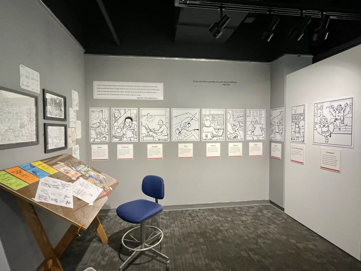 Art gallery corner with sketches on walls, an angled wooden desk with colorful papers, and a blue swivel chair on a gray floor.