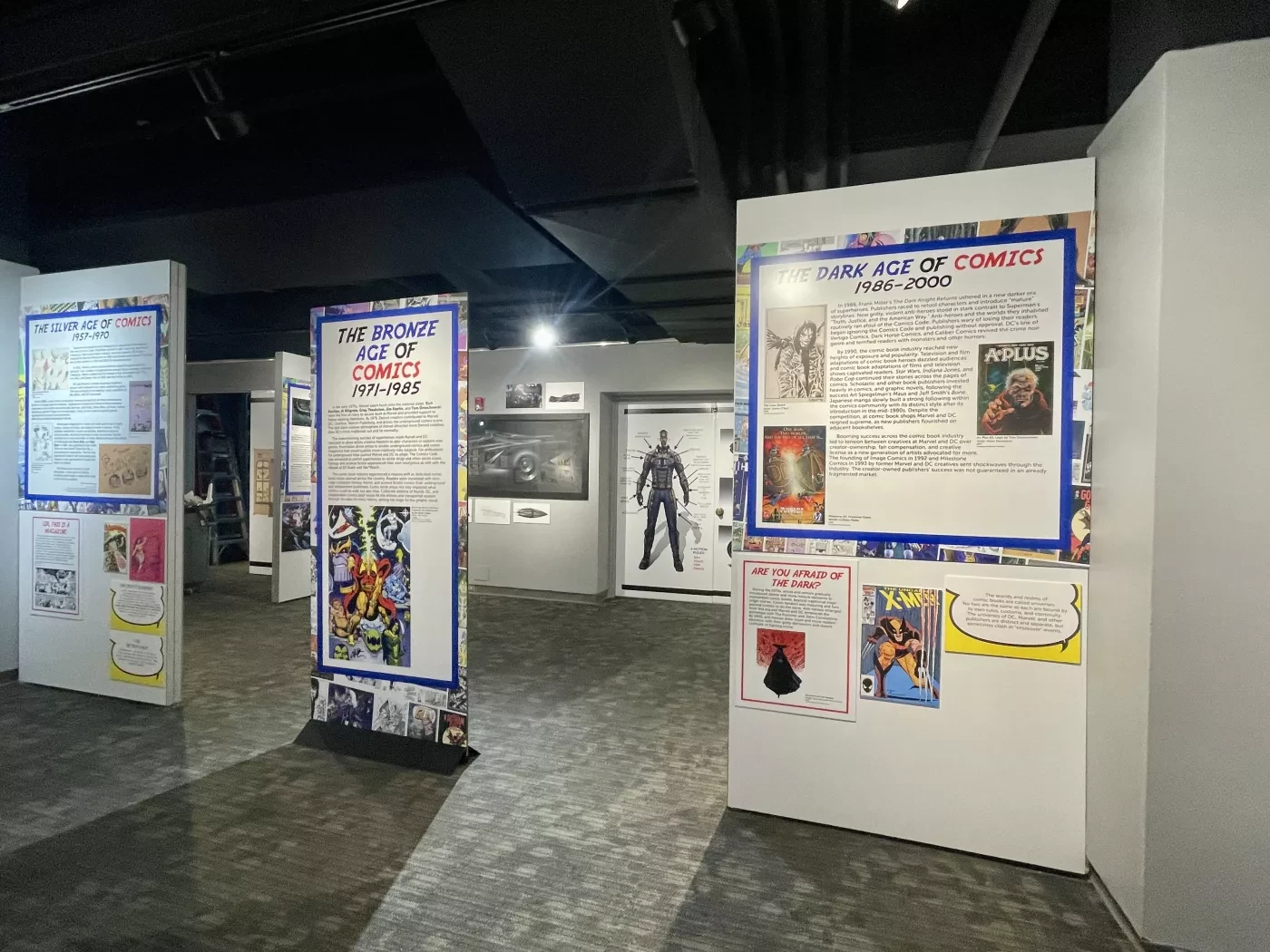 Museum exhibition room with comic displays on white panels, featuring colorful images and text under spotlights.