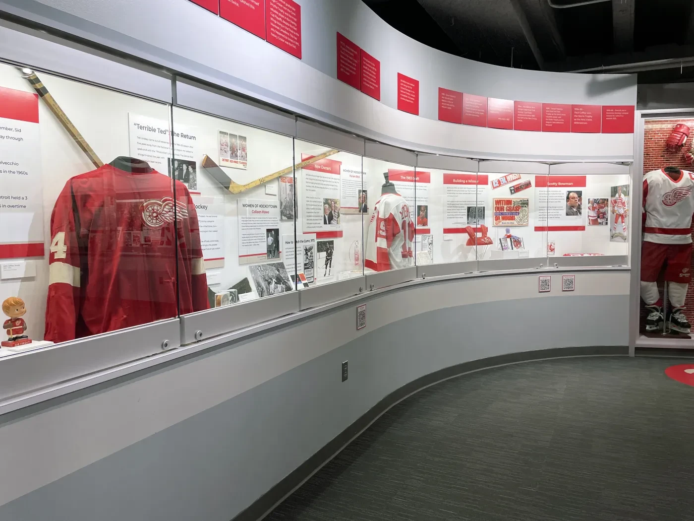Hockey museum display with jerseys, sticks, and memorabilia in a curved glass case, featuring mostly red and white colors.