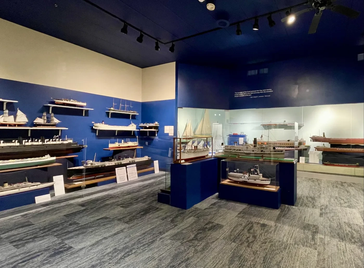 Museum exhibit room with model ships displayed on walls and in glass cases, under dim lighting.
