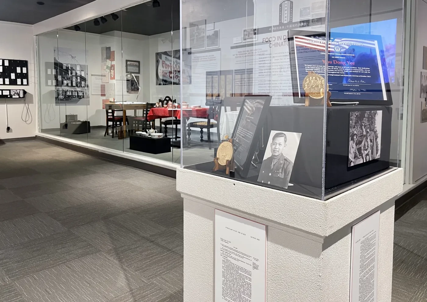 Museum exhibit with a glass display case showcasing awards, photographs, and memorabilia. Tables and historical images in the background.